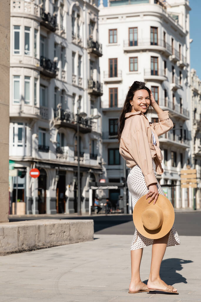 полная длина счастливой женщины в модной одежде держа соломенную шляпу на городской улице - Фото, изображение