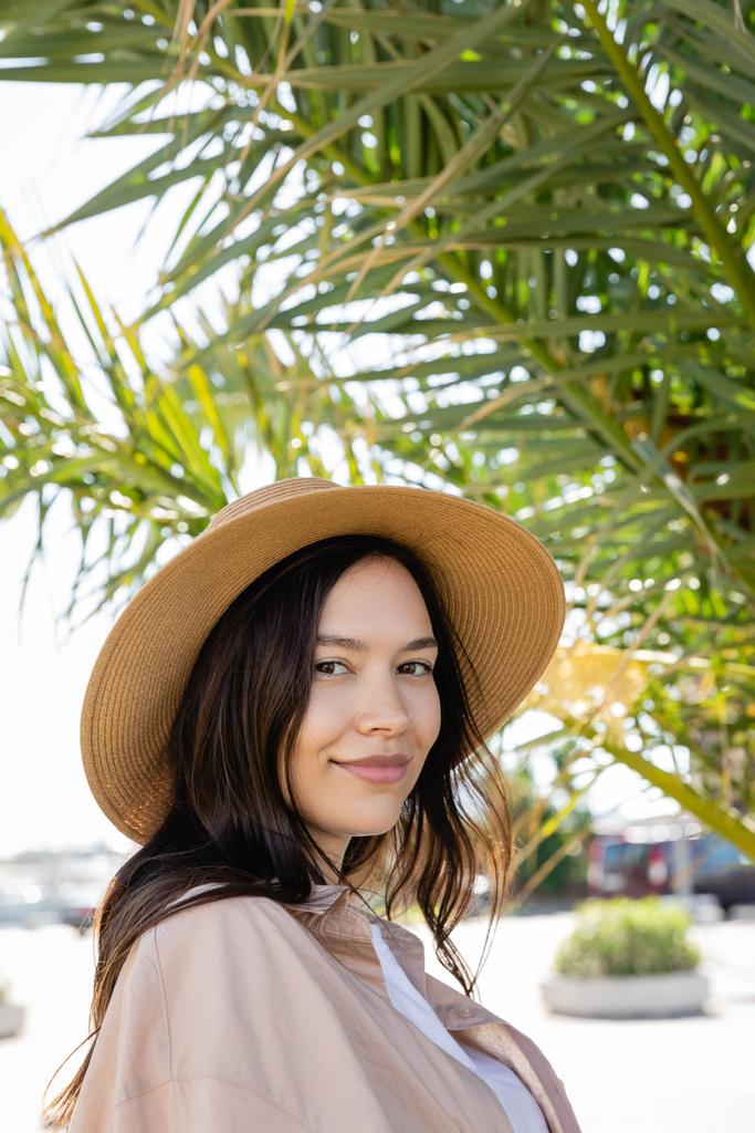 portrait of brunette woman in straw hat smiling at camera near blurred palm tree - Photo, image