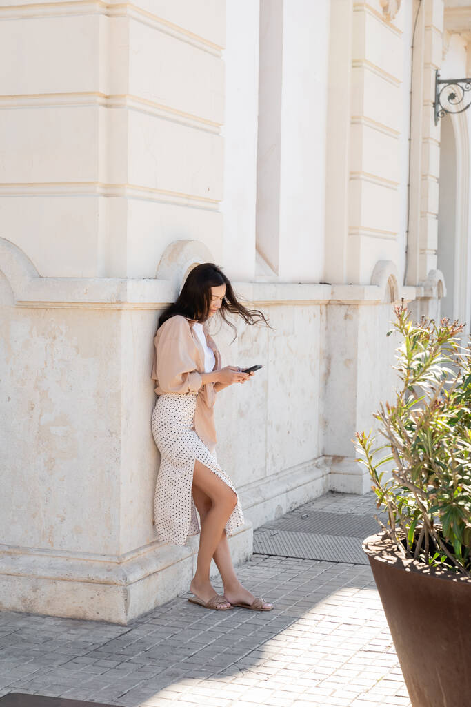 plný pohled na stylovou ženu stojící v blízkosti bílé budovy a pomocí smartphonu - Fotografie, Obrázek