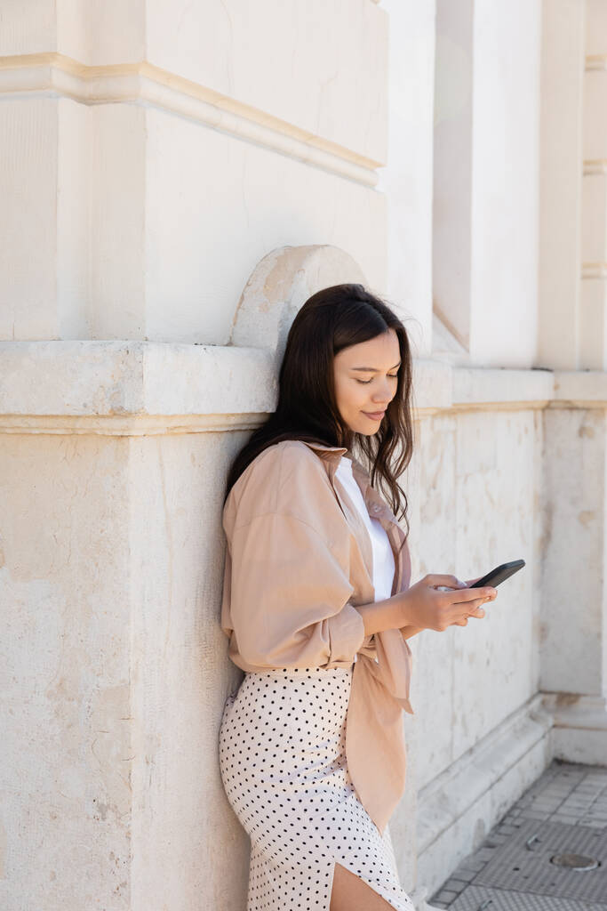 положительная брюнетка женщина в стильной одежде держа смартфон возле белой стены - Фото, изображение