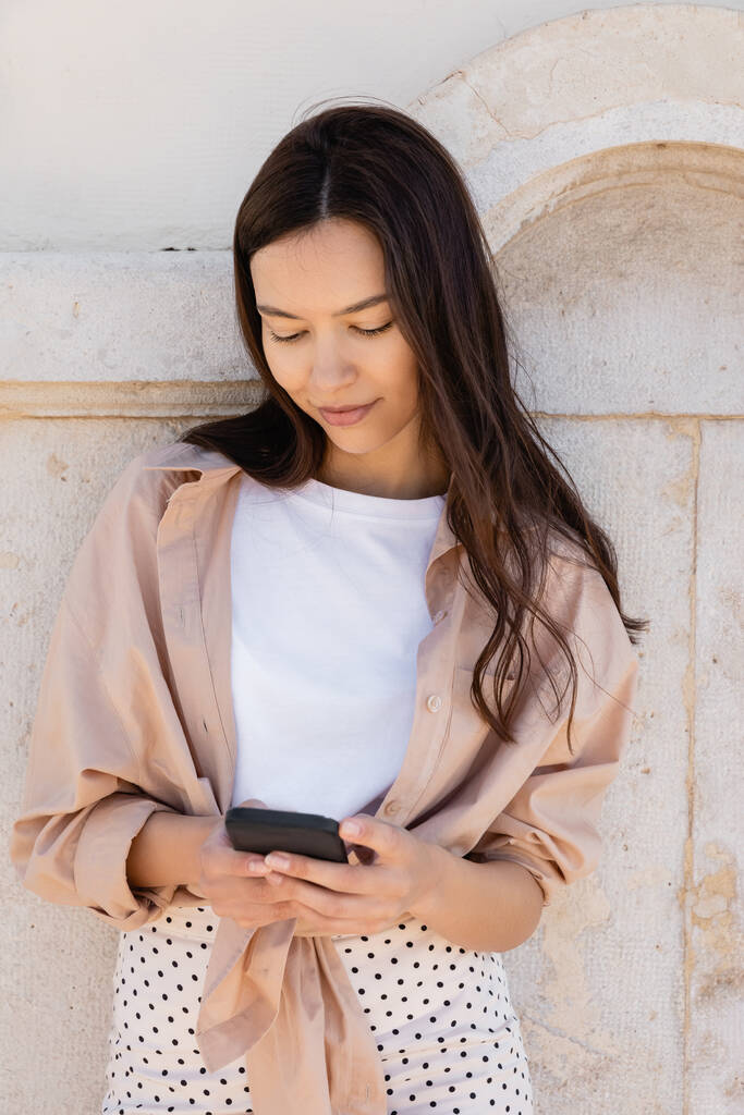 pozitivní brunetka žena stojící u bílé zdi a chatování na mobilním telefonu - Fotografie, Obrázek