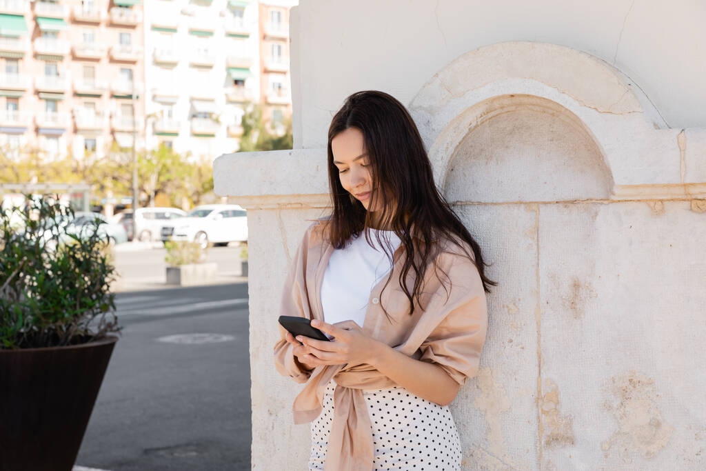 smiling woman in trendy clothes using mobile phone near white wall - Foto, Imagen