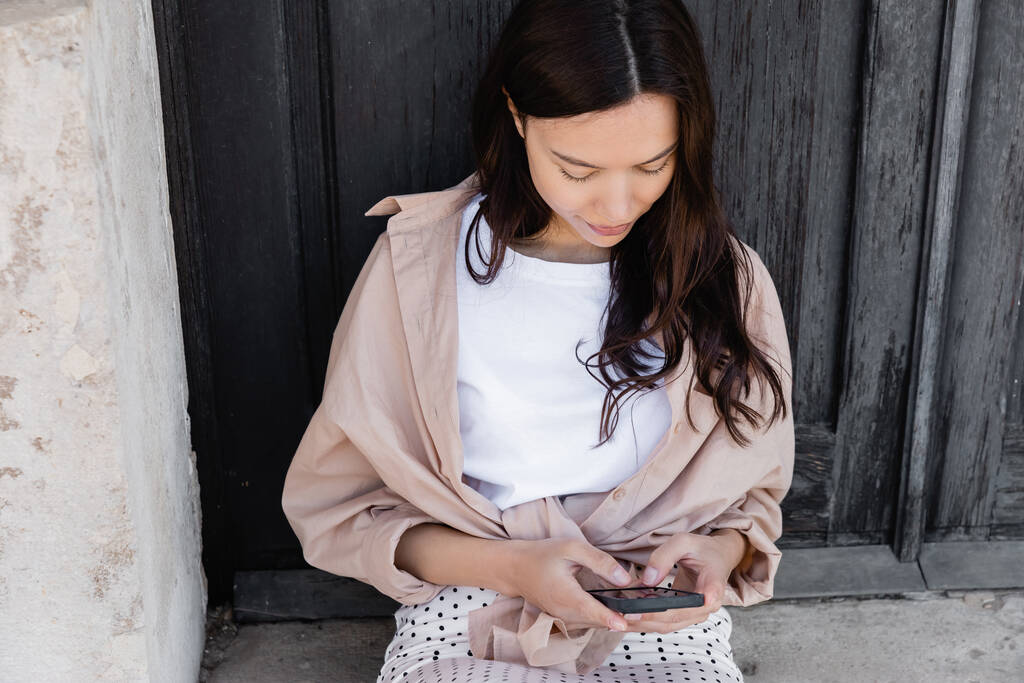 brunette woman sitting near black wooden door and messaging on smartphone - 写真・画像