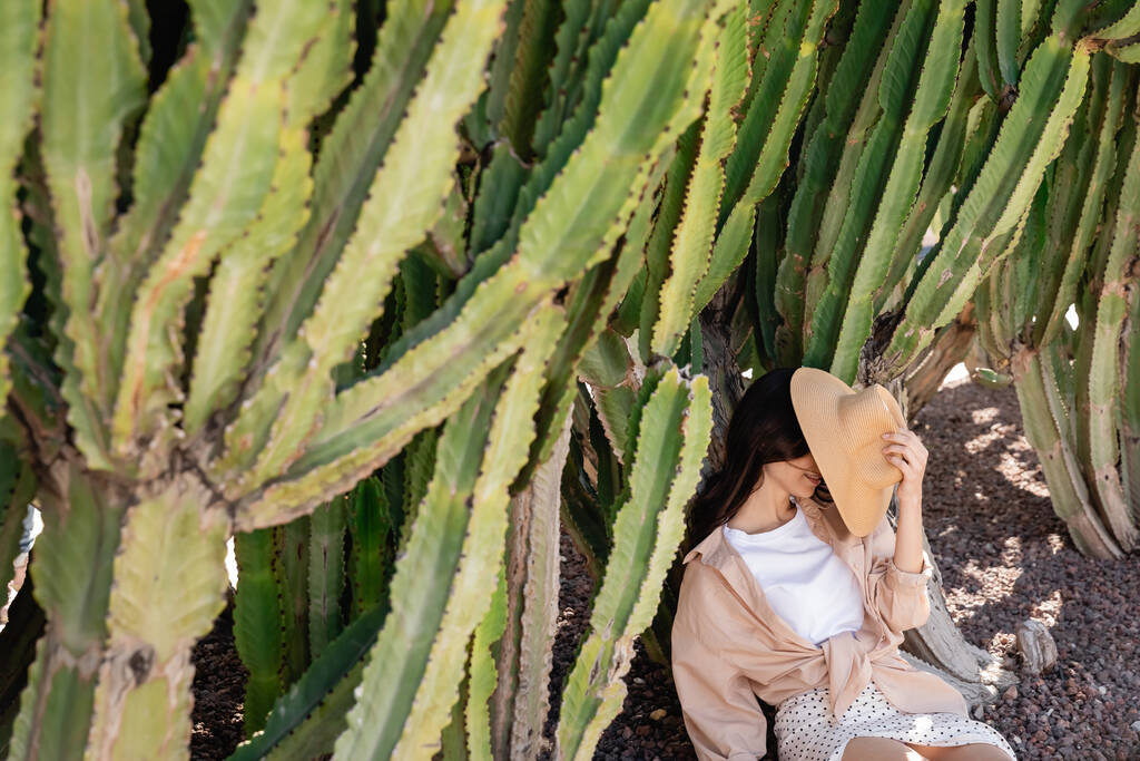 woman obscuring face with straw hat while sitting near giant cacti - Foto, Bild
