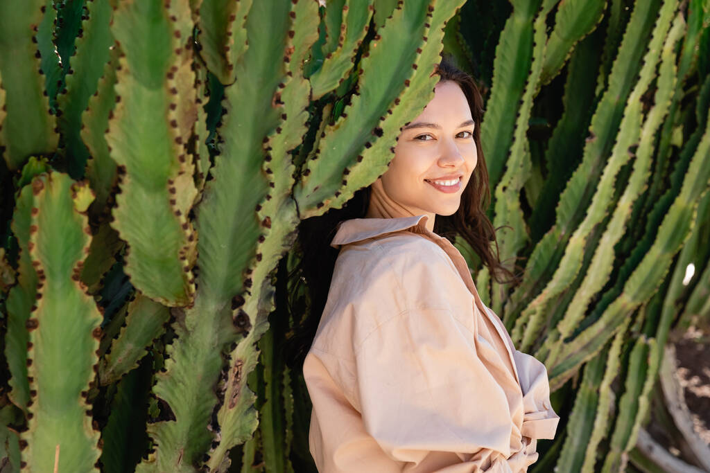 boldog nő bézs ing nézi kamera közelében zöld kaktuszok - Fotó, kép