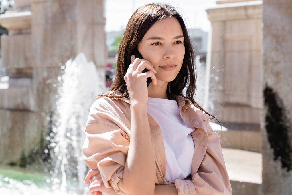 брюнетка жінка дивиться далеко під час розмови на мобільному телефоні біля розмитого фонтану
 - Фото, зображення