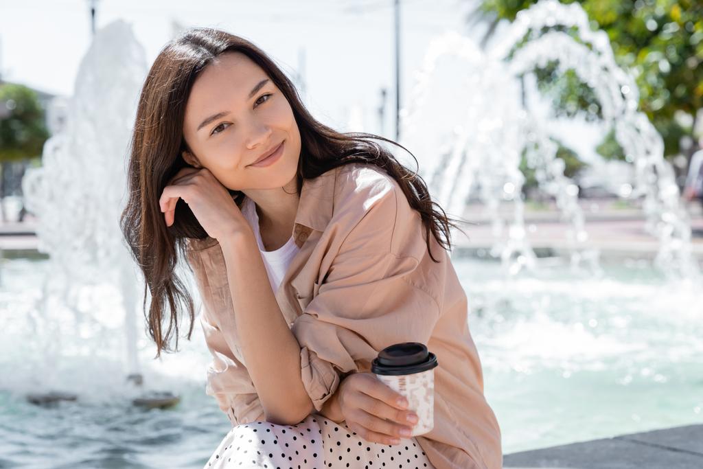 mulher sorrindo em camisa bege segurando café para ir e sorrindo para a câmera perto de fonte turva - Foto, Imagem