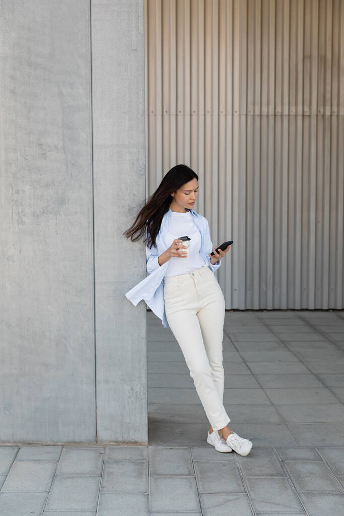 longitud completa de la mujer morena en pantalones blancos de pie con teléfono celular y taza de papel cerca de la pared gris - Foto, imagen