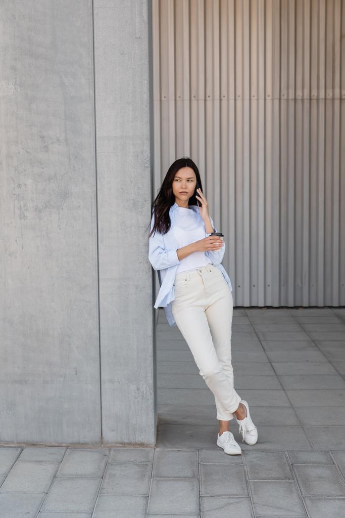full length of woman in white pants holding paper cup and talking on smartphone near grey wall - Foto, Imagem