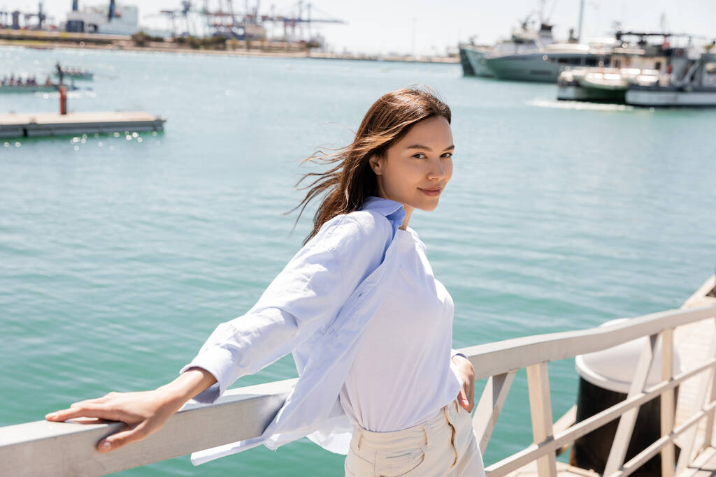 mujer sonriente en camisa azul mirando a la cámara mientras está de pie en el muelle en el puerto marítimo - Foto, imagen