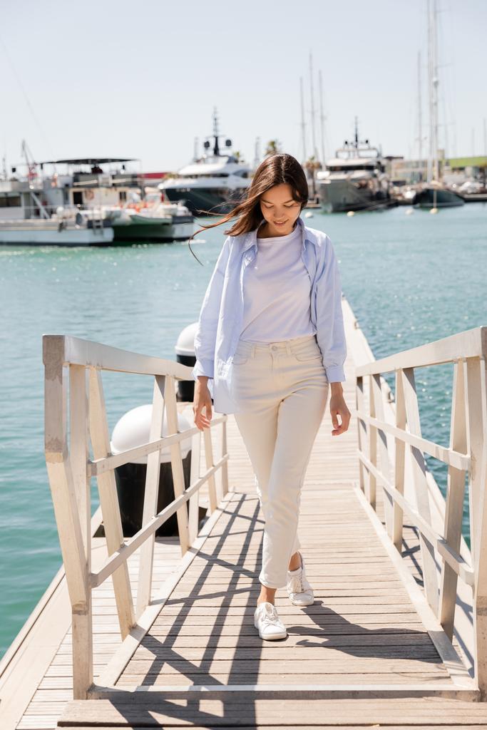 volle Länge der lächelnden Frau in blauem Hemd und weißer Hose, die auf der Seebrücke im Seehafen spaziert - Foto, Bild
