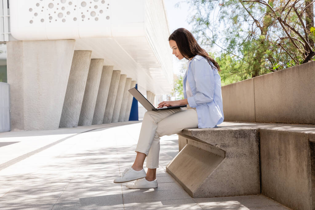 side view of brunette woman in blue shirt and white pants using laptop outdoors - Photo, Image