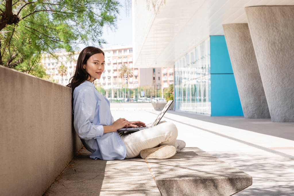 brunette woman in blue shirt sitting with laptop on street and looking at camera - Foto, Imagem
