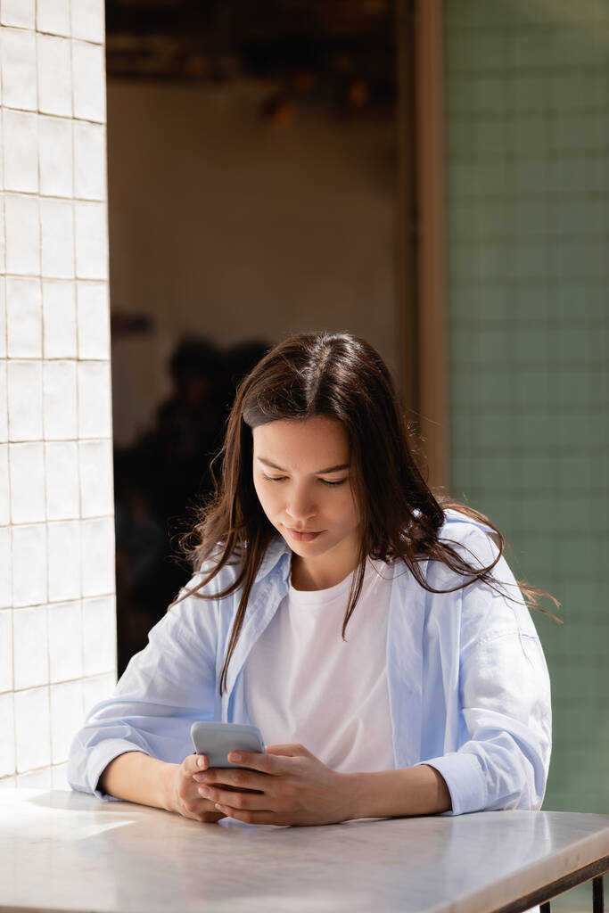 brunette nainen sininen paita messaging älypuhelimella kahvilan terassilla - Valokuva, kuva