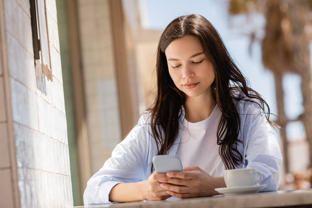 brunette woman using smartphone near coffee cup on summer terrace - Foto, immagini