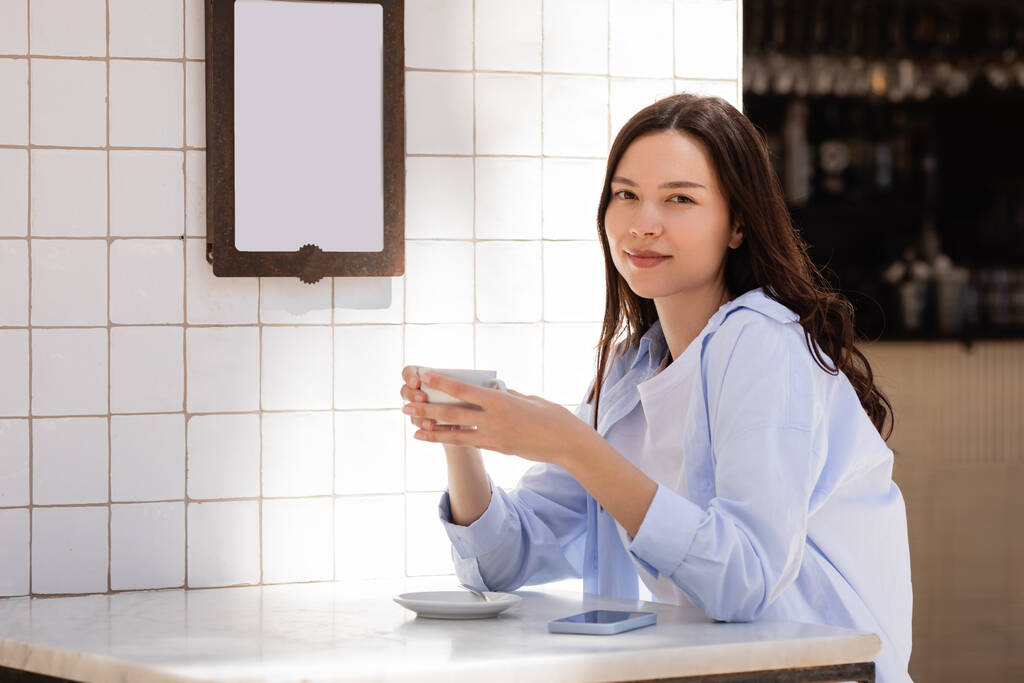 brünette Frau mit Tasse Kaffee blickt auf Caféterrasse in die Kamera - Foto, Bild