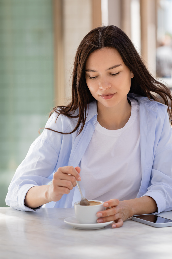 brunette woman holding teaspoon near coffee cup and smartphone with blank screen on cafe terrace - Foto, afbeelding