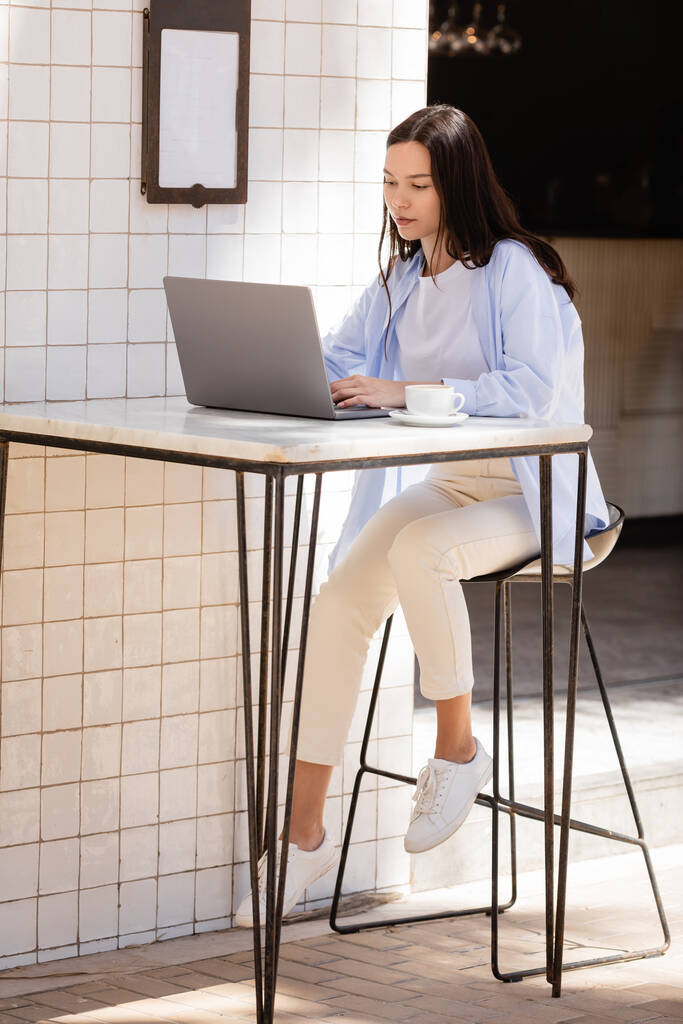 Вид брюнетки в полный рост, сидящей на ноутбуке возле чашки кофе на террасе кафе - Фото, изображение