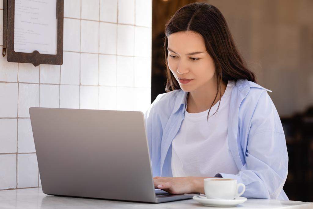 woman in blue shirt typing on laptop near coffee cup on cafe terrace - Zdjęcie, obraz