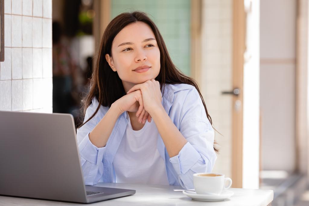 dreamy and positive woman looking away near coffee cup and laptop - Foto, afbeelding