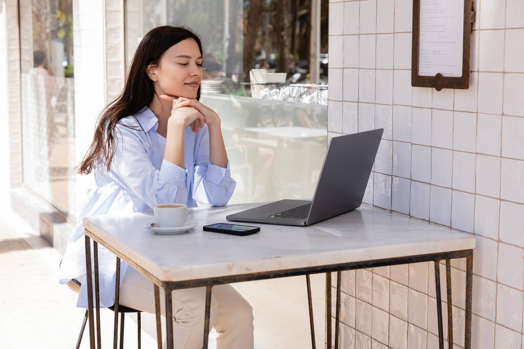 Lächelnde Frau blickt auf Laptop neben Smartphone und Kaffeetasse auf Sommerterrasse  - Foto, Bild