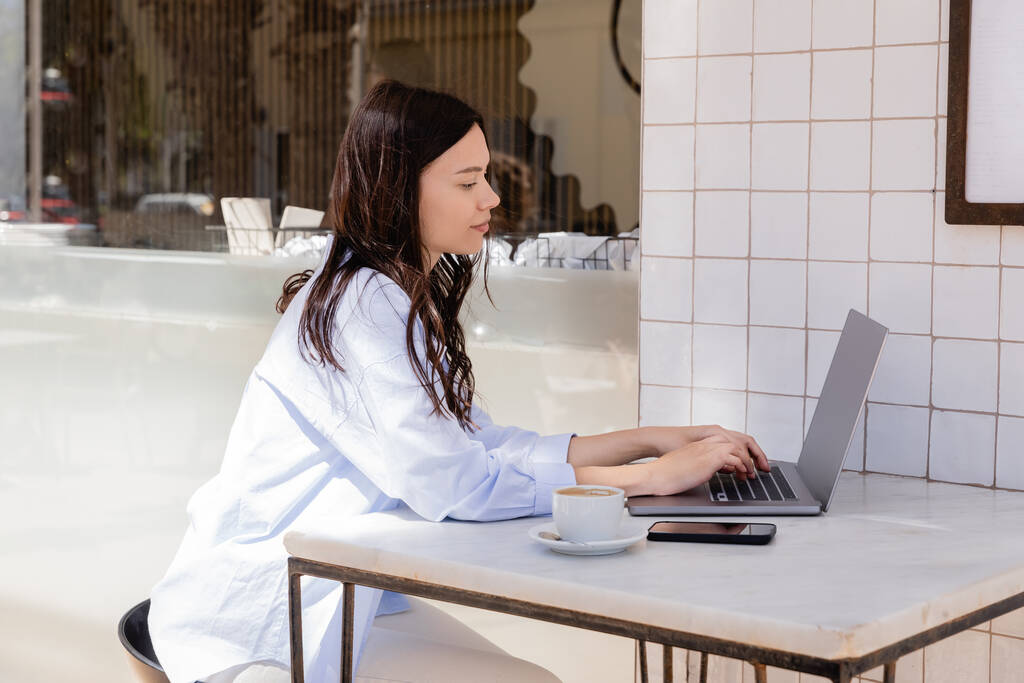 вид збоку жінки, що друкує на ноутбуці біля мобільного телефону та чашки кави у вуличному кафе
 - Фото, зображення