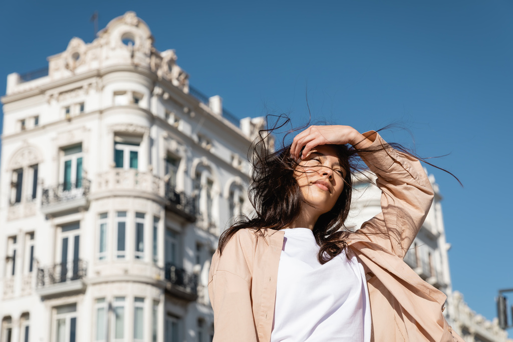 laag hoek uitzicht van brunette vrouw fixeren haar terwijl staan op de wind in de stad - Foto, afbeelding