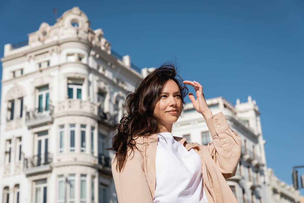 brunette woman standing on windy street near white buildings on blurred background - Photo, Image