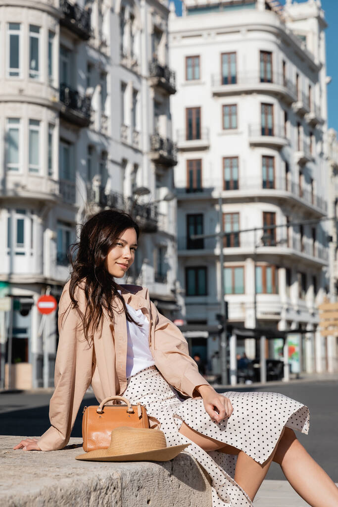 стильна брюнетка жінка сидить на вулиці біля солом'яного капелюха і сумочки
 - Фото, зображення