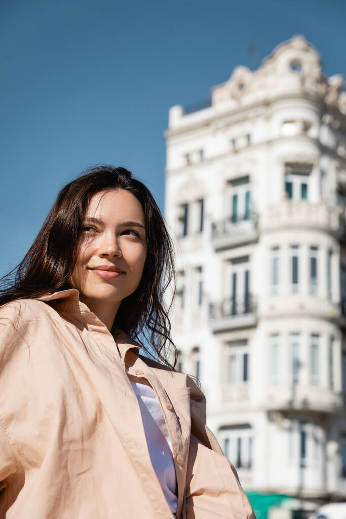 brunette woman in beige shirt smiling near white building on blurred background - Photo, Image