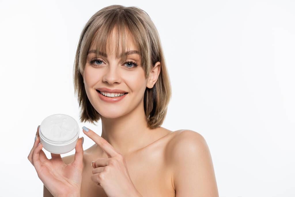 happy young woman with bangs pointing with finger at container with face cream isolated on white  - Photo, Image