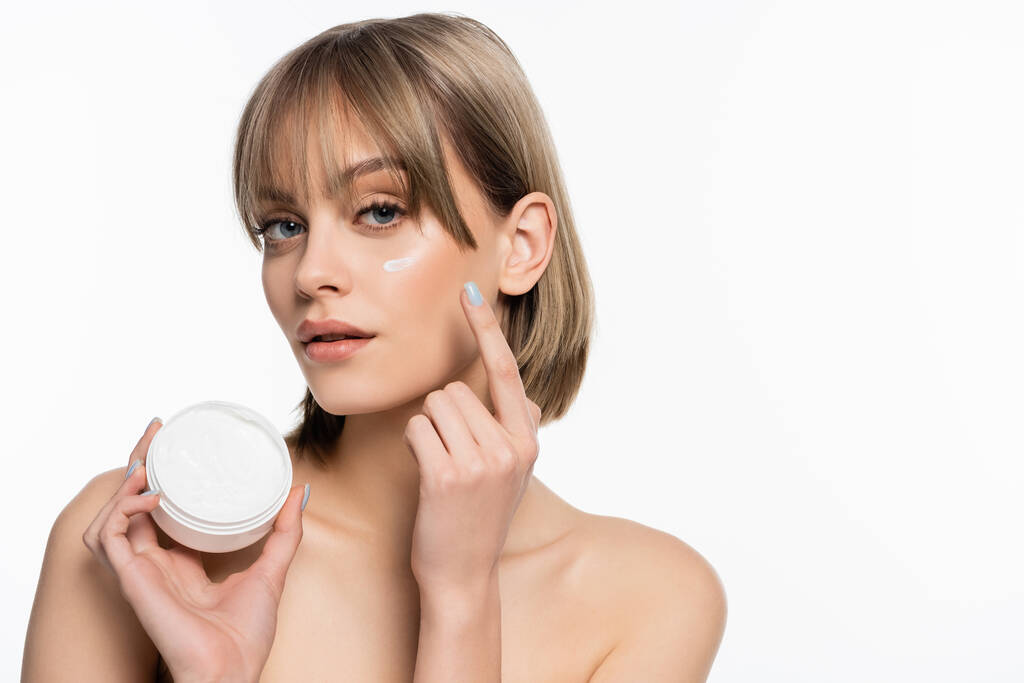 young woman with bangs and cream on cheek holding container isolated on white  - Foto, afbeelding