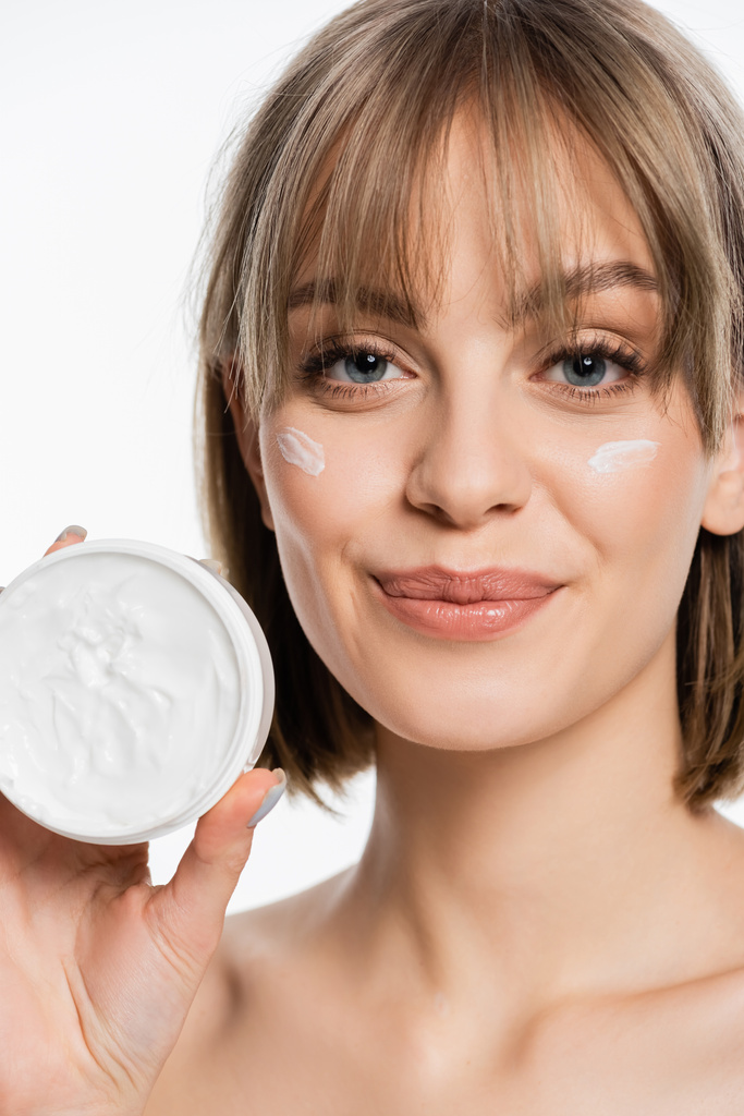 smiling woman with bangs and cream on face holding container while looking at camera isolated on white - Foto, Imagem
