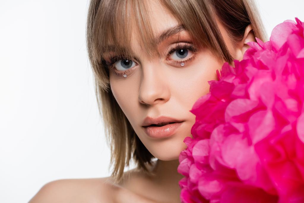 young woman with bangs and rhinestones under blue eyes near pink flower isolated on white - Foto, Bild