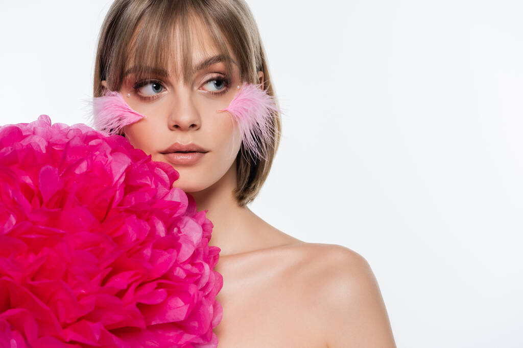 mujer bonita con elementos decorativos en el maquillaje y plumas de color rosa en las mejillas cerca de flor brillante aislado en blanco - Foto, Imagen