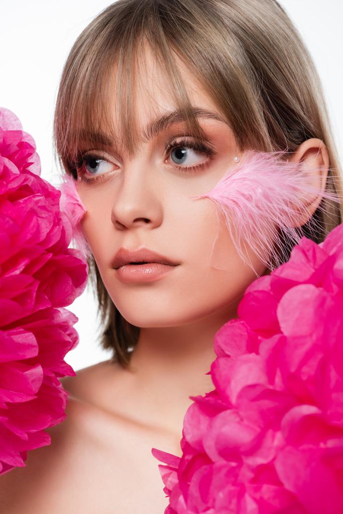 young woman with decorative elements in makeup and pink feathers on cheeks near flowers isolated on white - Fotografie, Obrázek