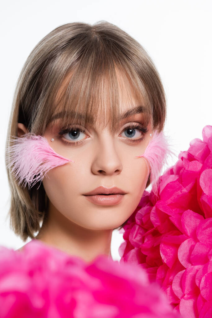 młoda kobieta z elementami dekoracyjnymi w makijażu, grzywki i różowe pióra na policzkach w pobliżu kwiatów odizolowanych na białym - Zdjęcie, obraz