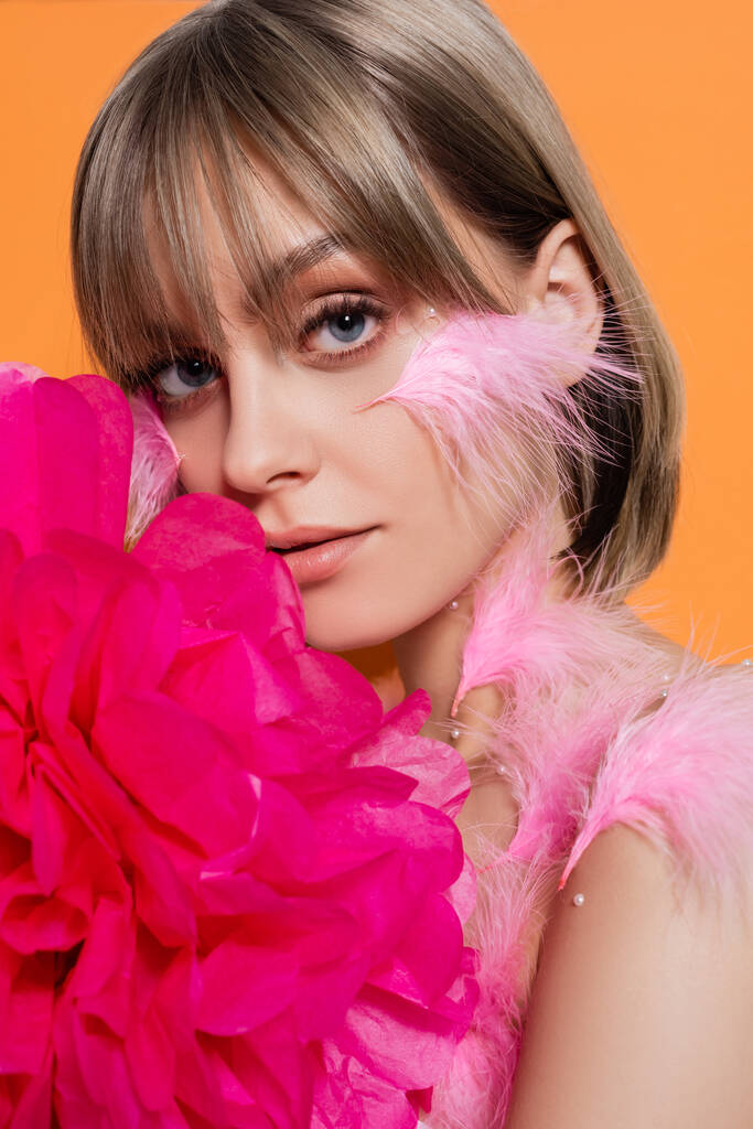 young woman with decorative beads in makeup and feathers on cheeks near pink flower isolated on orange - Фото, изображение