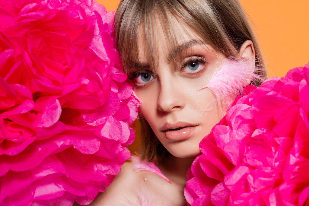 young woman with decorative beads in makeup and feathers on cheeks near pink flowers isolated on orange - Photo, image