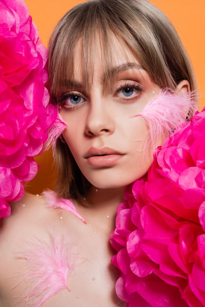 woman with decorative beads in makeup and pink feathers on cheeks near flowers isolated on orange - Photo, image