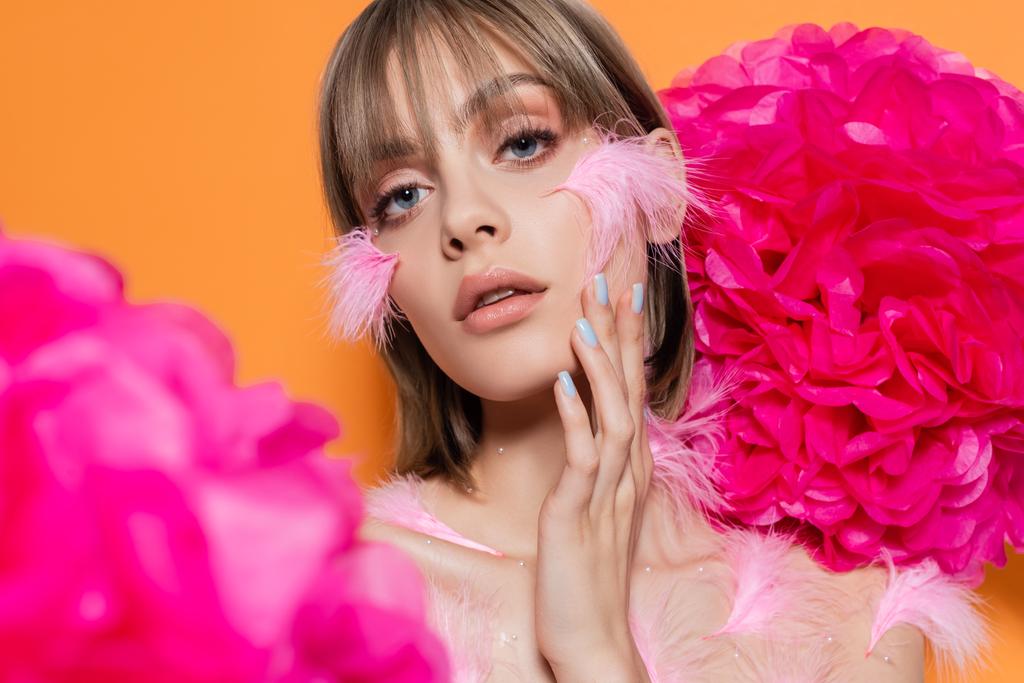 молода жінка з декоративними елементами в макіяжі і рожевим пір'ям на щоках біля квітів ізольовані на апельсині
 - Фото, зображення