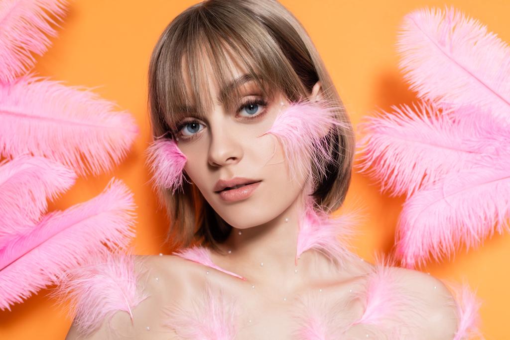 young woman with decorative beads in makeup near pink feathers isolated on orange - Foto, afbeelding