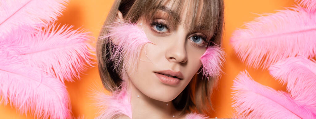 young woman with decorative beads in makeup near pink feathers isolated on orange, banner - 写真・画像