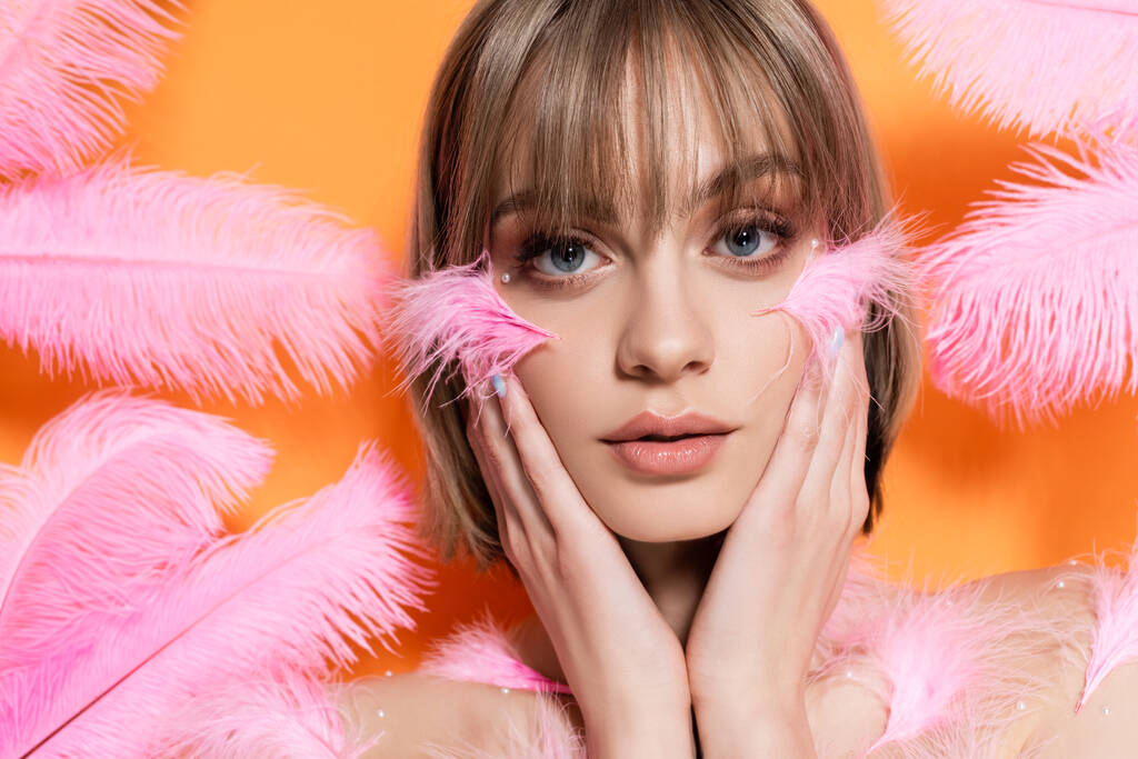 young woman with decorative beads in makeup touching face near pink feathers isolated on orange - Фото, изображение