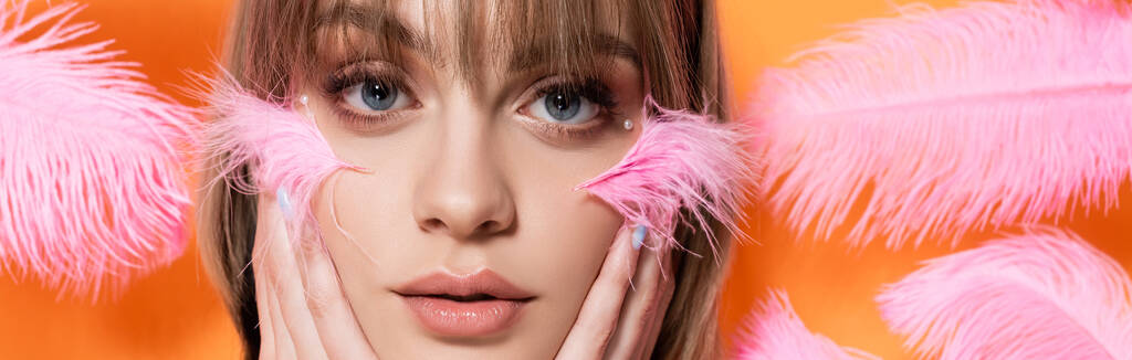 young woman with decorative beads in makeup touching face near pink feathers isolated on orange, banner - Fotografie, Obrázek