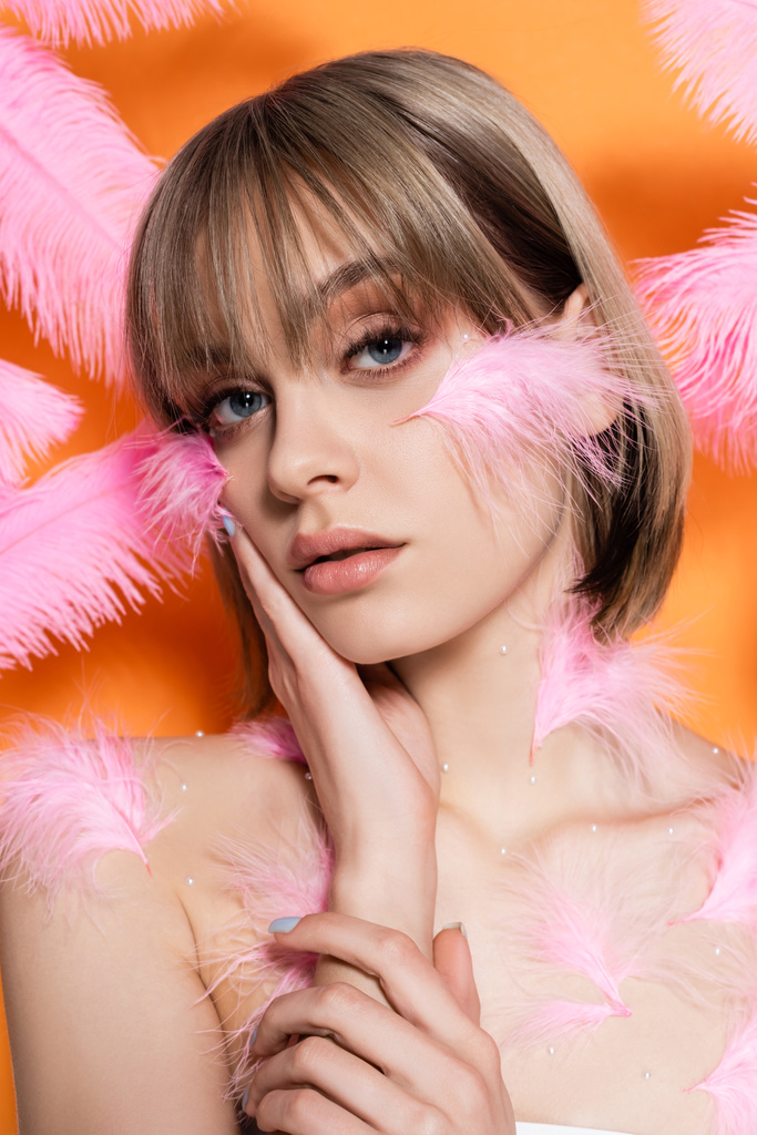 メイクは装飾ビーズを持つ若い女性オレンジに隔離されたピンクの羽の近くに頬に触れる - 写真・画像