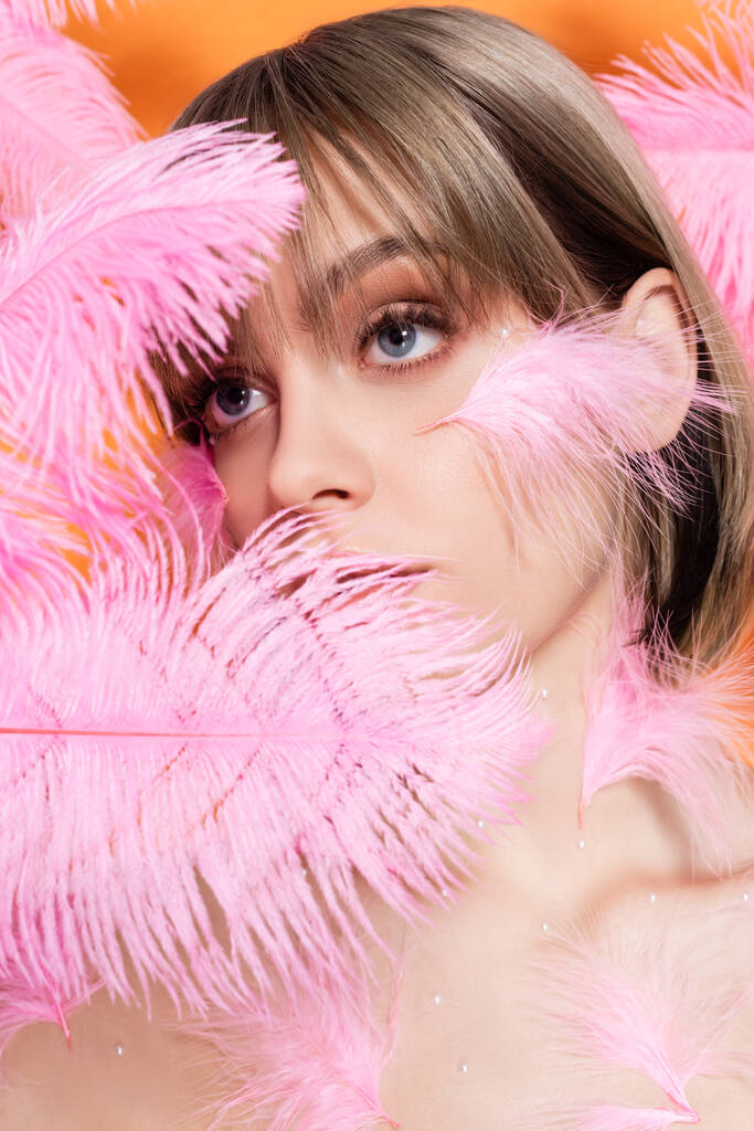 young woman with decorative beads in makeup looking away through pink feathers isolated on orange - Foto, Bild