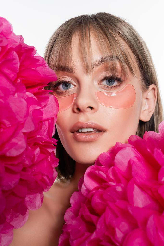 Nuori nainen hydrogeeli silmä laikkuja ja siniset silmät lähellä kirkkaan vaaleanpunaisia kukkia eristetty valkoinen - Valokuva, kuva