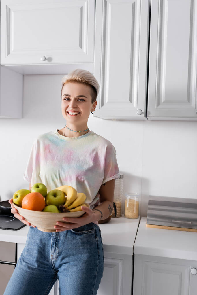 cheerful and stylish woman with bowl of ripe fruits smiling at camera in kitchen - Foto, afbeelding