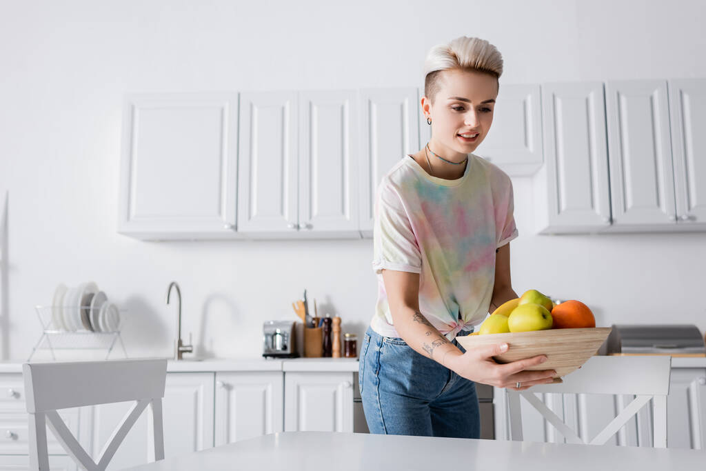 mulher positiva em t-shirt segurando tigela com frutas frescas na cozinha - Foto, Imagem